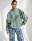 Woman wearing the moss green God Is Greater Hoodie with jeans, showing the front kangaroo pocket.