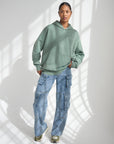 Full-length view of a woman styling the God Is Greater Moss Hoodie with loose-fitting jeans.