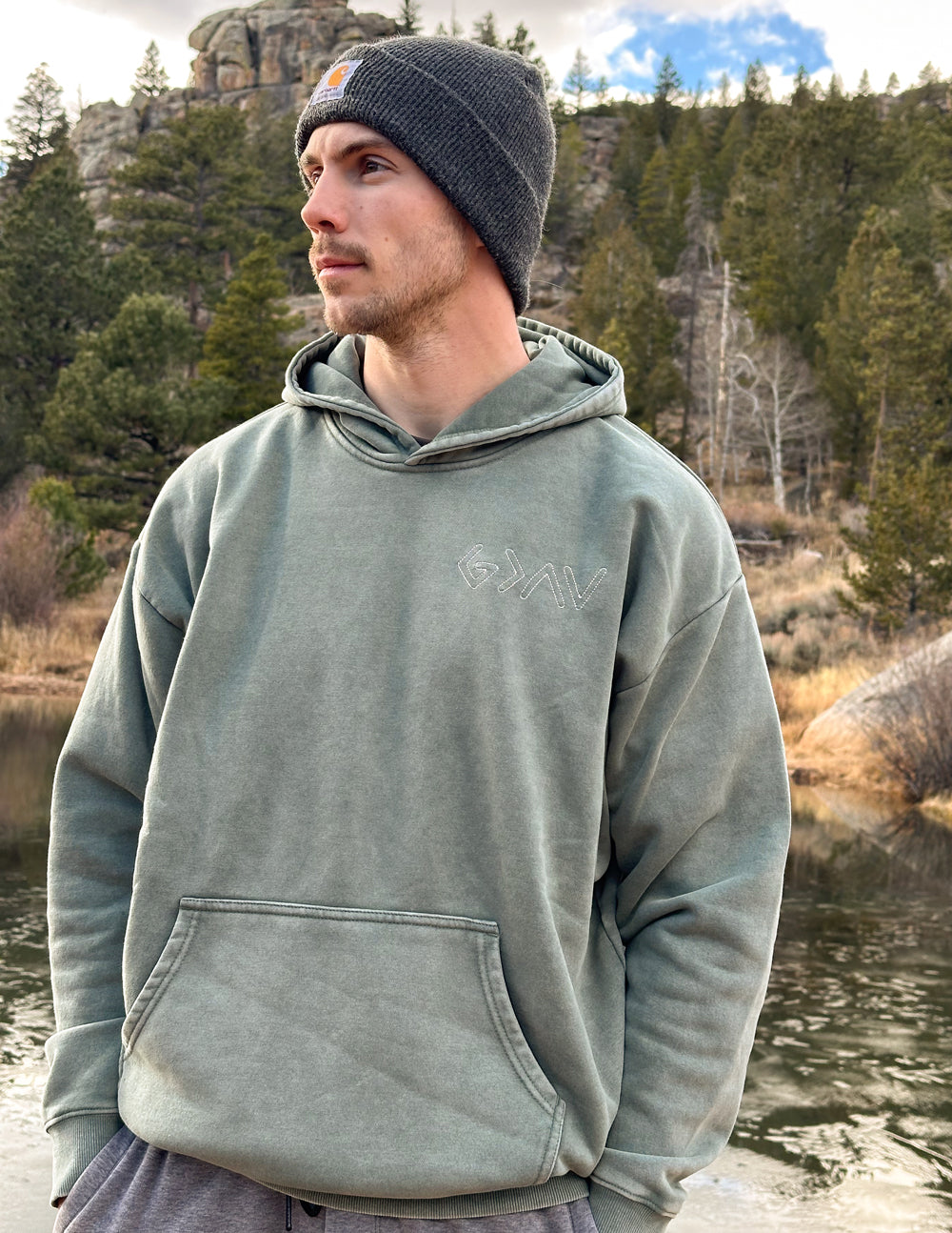 Man wearing the God Is Greater Moss Hoodie with a dark beanie outdoors.