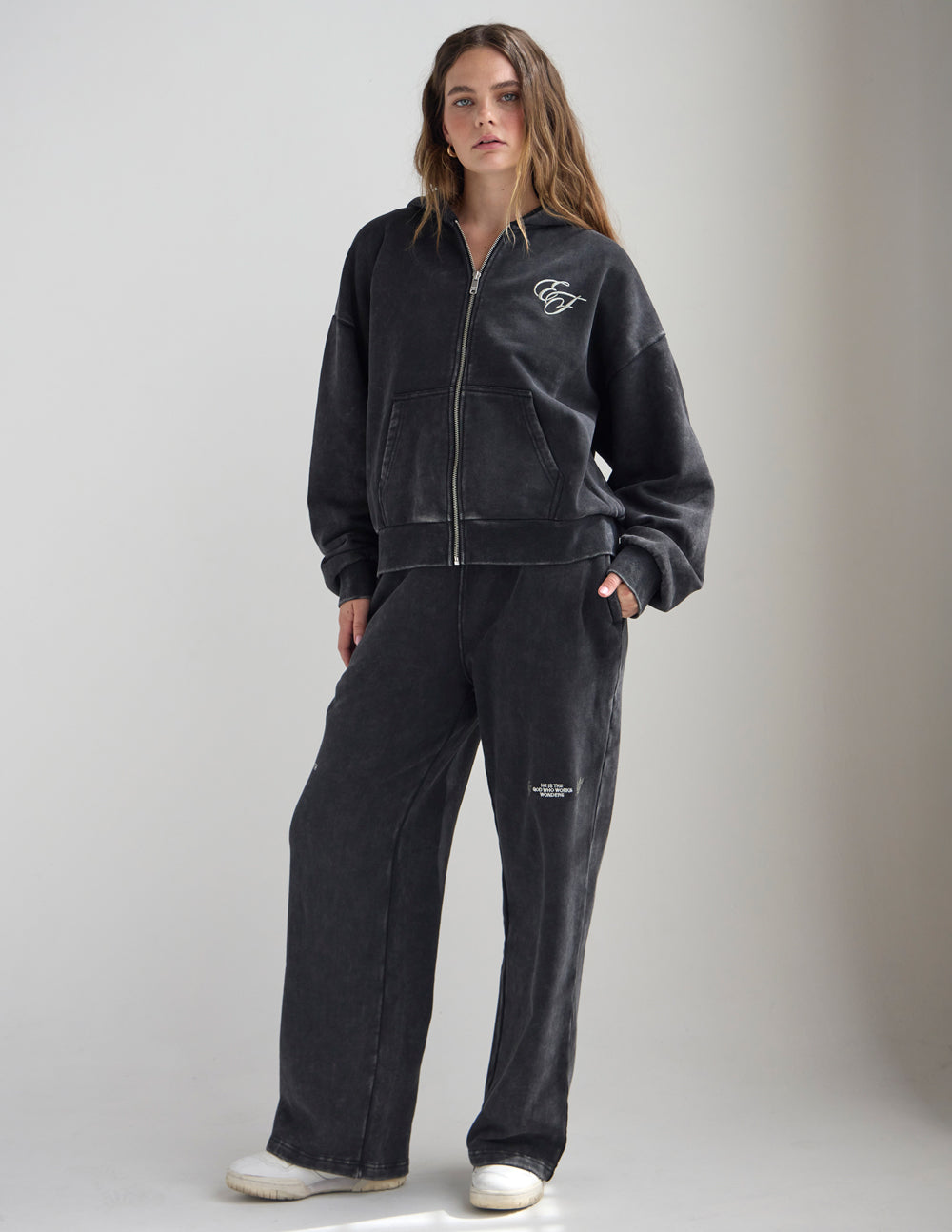 Full-length view of a woman wearing the black God of Miracles zip hoodie with coordinating pants.