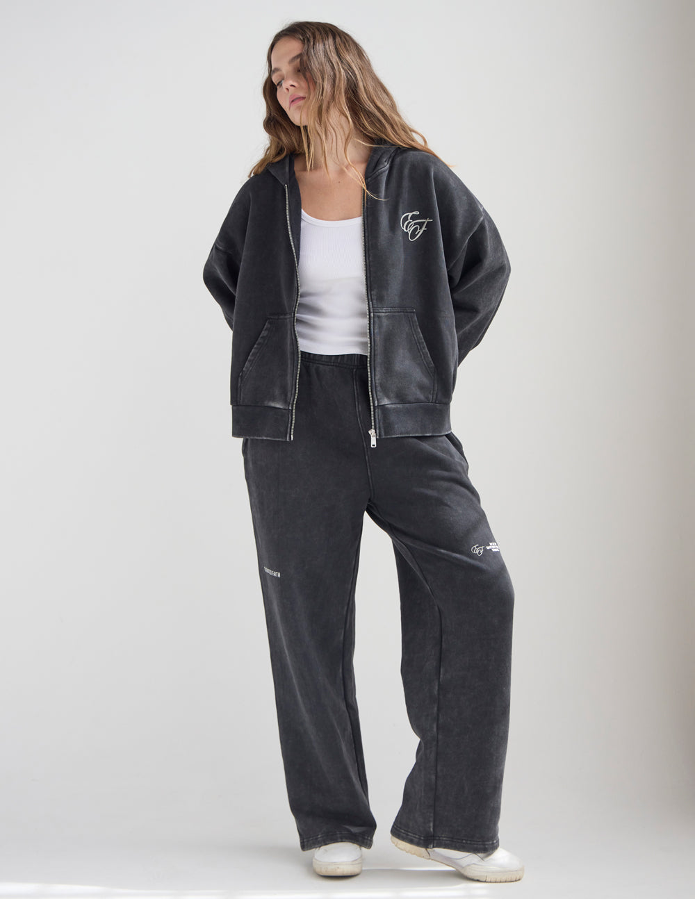 Woman wearing the black God of Miracles Hoodie open over a white top with coordinating pants.