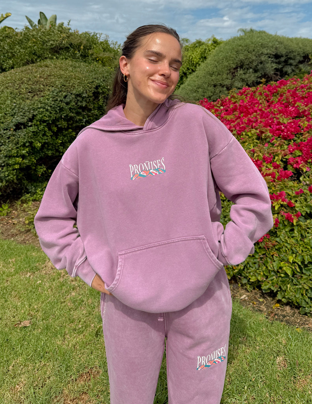 Woman wearing the Bordeaux God’s Promises Hoodie with a front kangaroo pocket outdoors beside flowering shrubs.