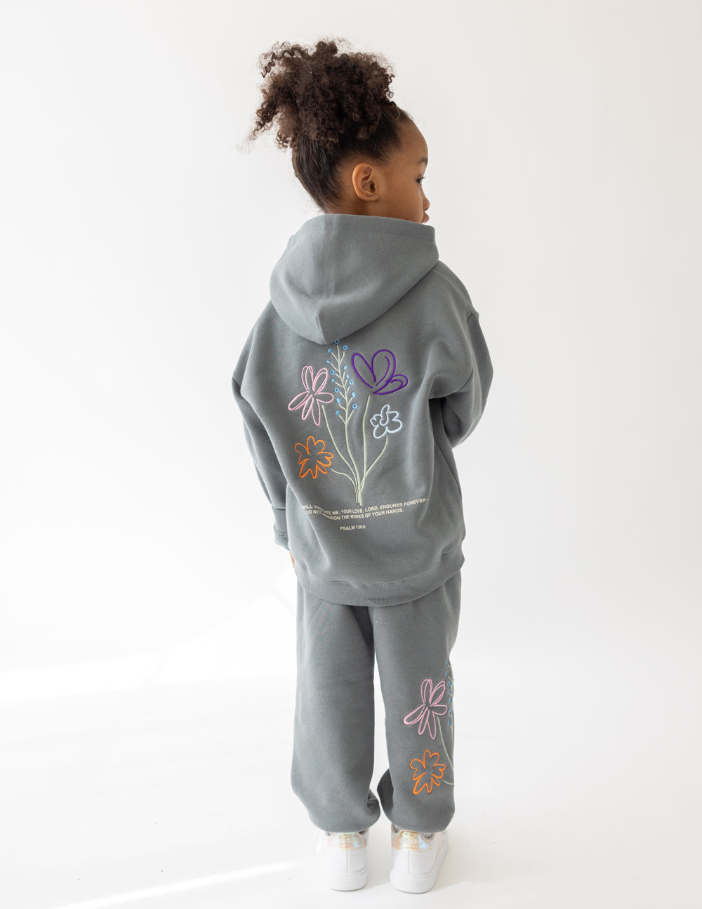 Back view of a child wearing the God’s Still Working Sedona Sage hoodie with colorful floral artwork.