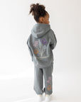 Back view of a child wearing the God’s Still Working Sedona Sage hoodie with colorful floral artwork.