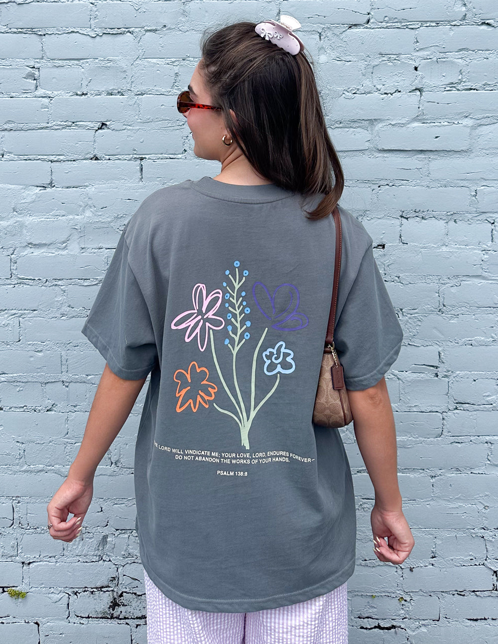 Back of the God’s Still Working Sedona Sage tee featuring colorful floral artwork against a pale blue brick wall