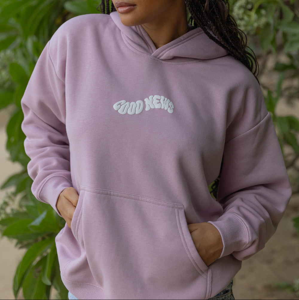 Person wearing a light purple hoodie with 'Good News' text, standing outdoors with greenery in the background