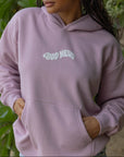 Person wearing a light purple hoodie with 'Good News' text, standing outdoors with greenery in the background