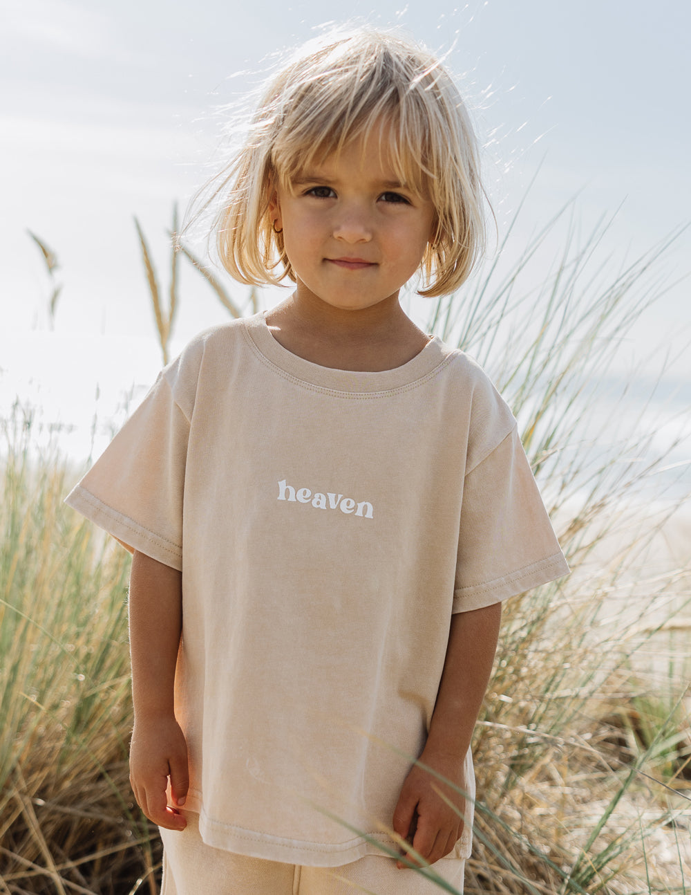 Child wearing the Heaven Kids Tee in Nomad with white heaven lettering, standing near beach grass.