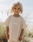 Child wearing the Heaven Kids Tee in Nomad with white heaven lettering, standing near beach grass.