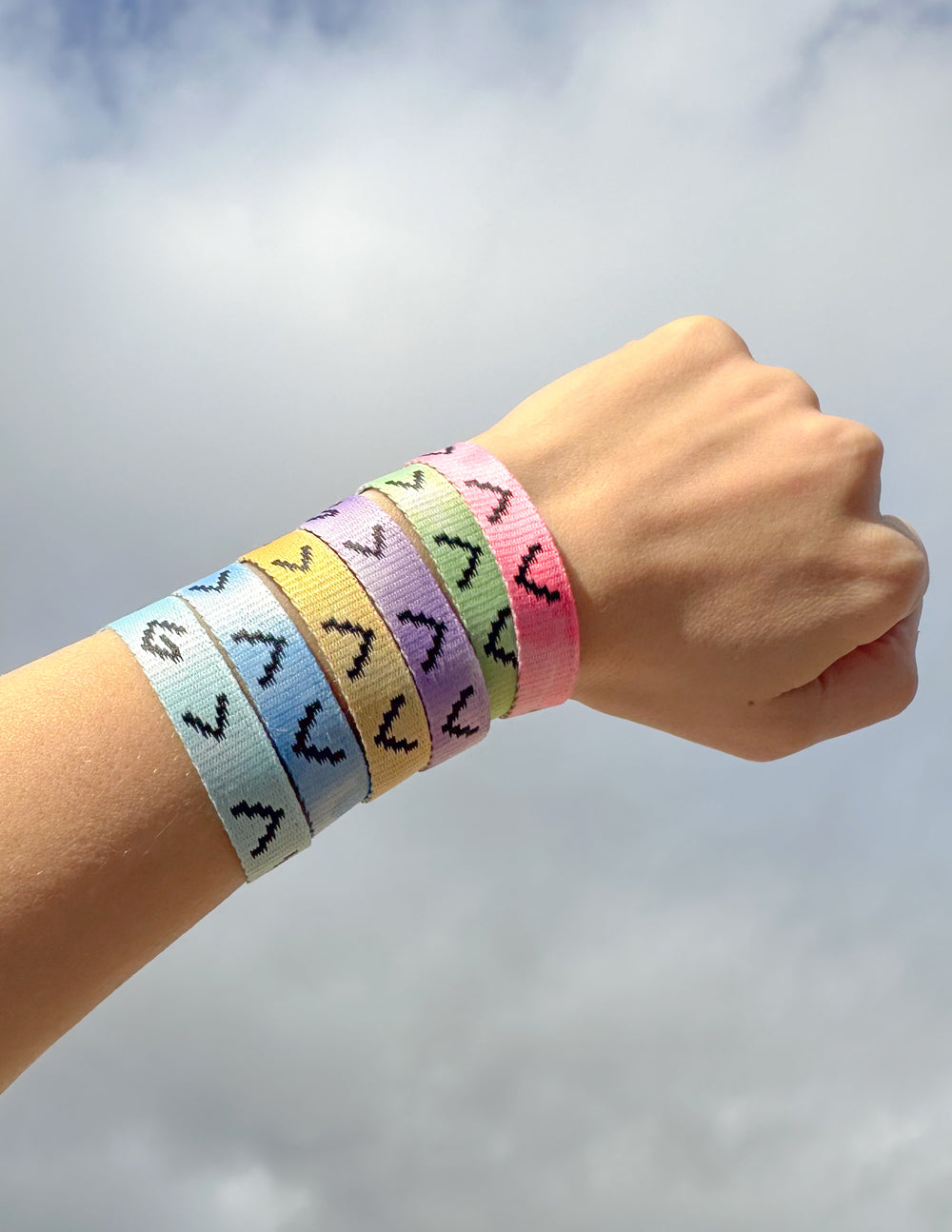 Six colorful Highs and Lows tie-dye bracelets stacked on a wrist against a cloudy sky