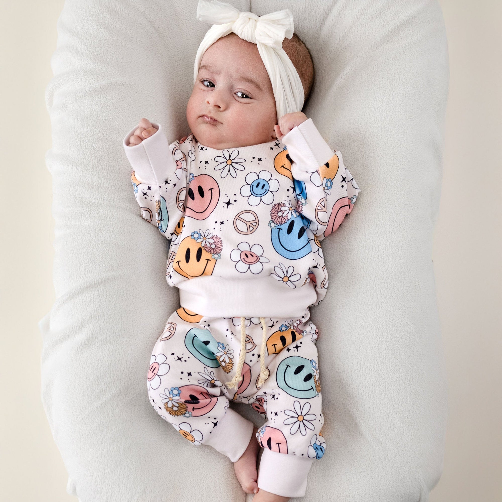 Baby wearing a colorful outfit with smiley faces and patterns, lying on a white cushion.