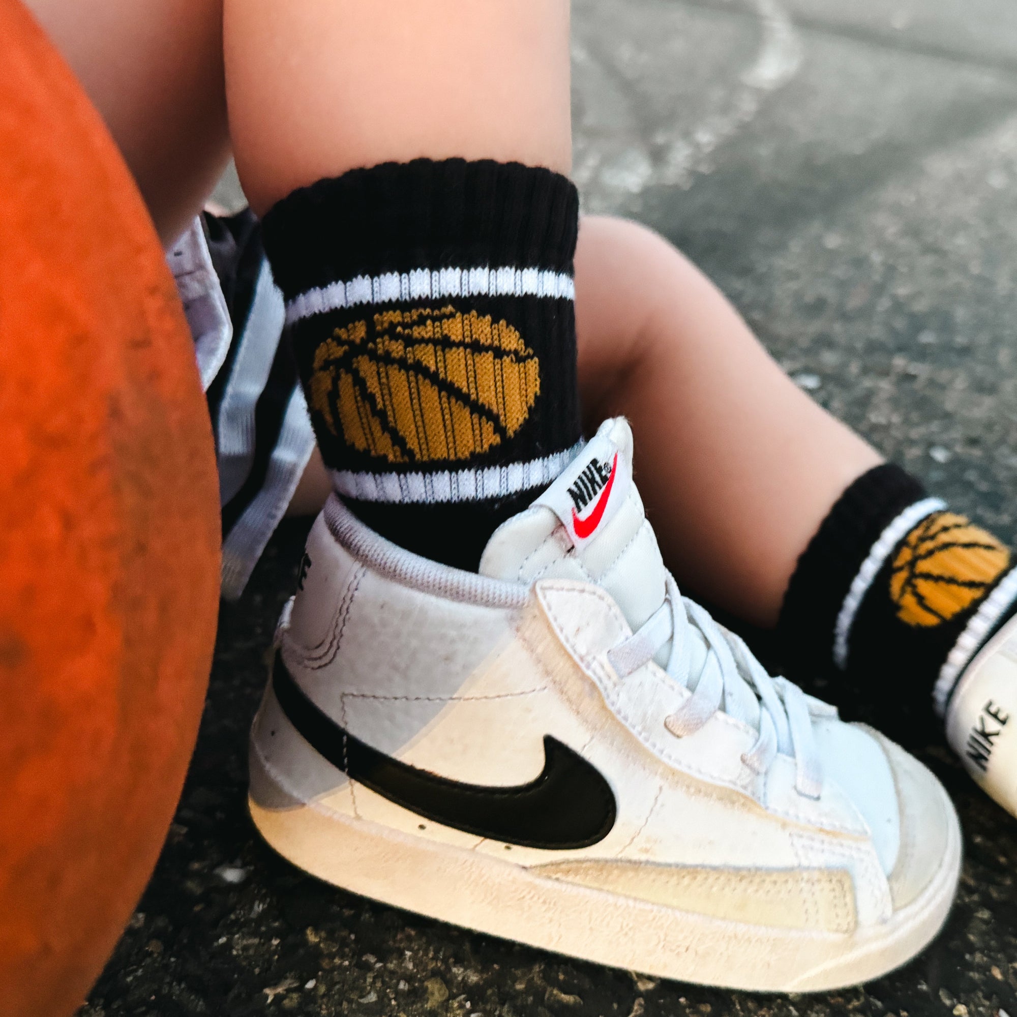 Person wearing white sneakers with black swoosh logo, black socks with basketball design, and orange ball on a dark surface.