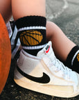 Person wearing white sneakers with black swoosh logo, black socks with basketball design, and orange ball on a dark surface.