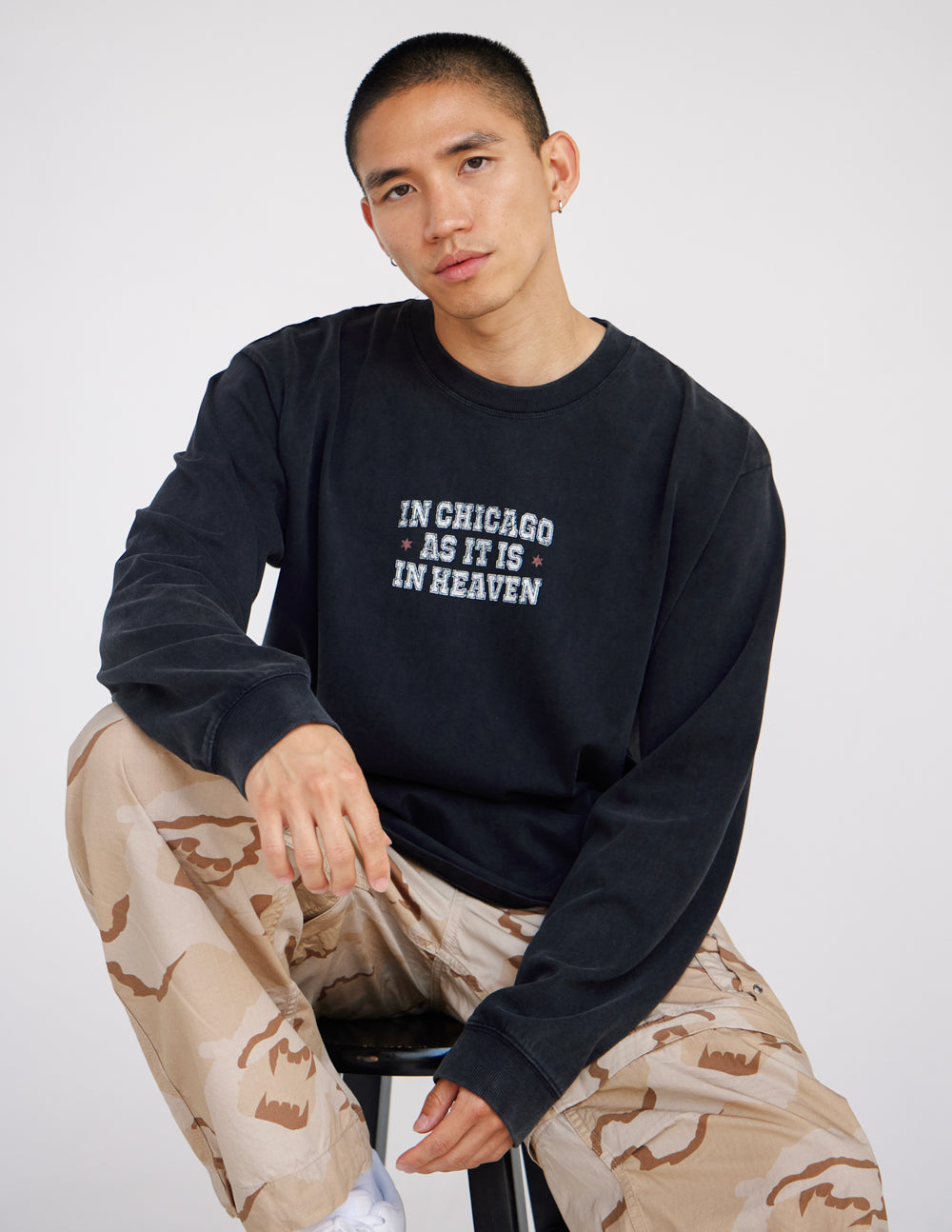 Seated man wearing black In Chicago as It Is in Heaven long sleeve tee with camouflage pants