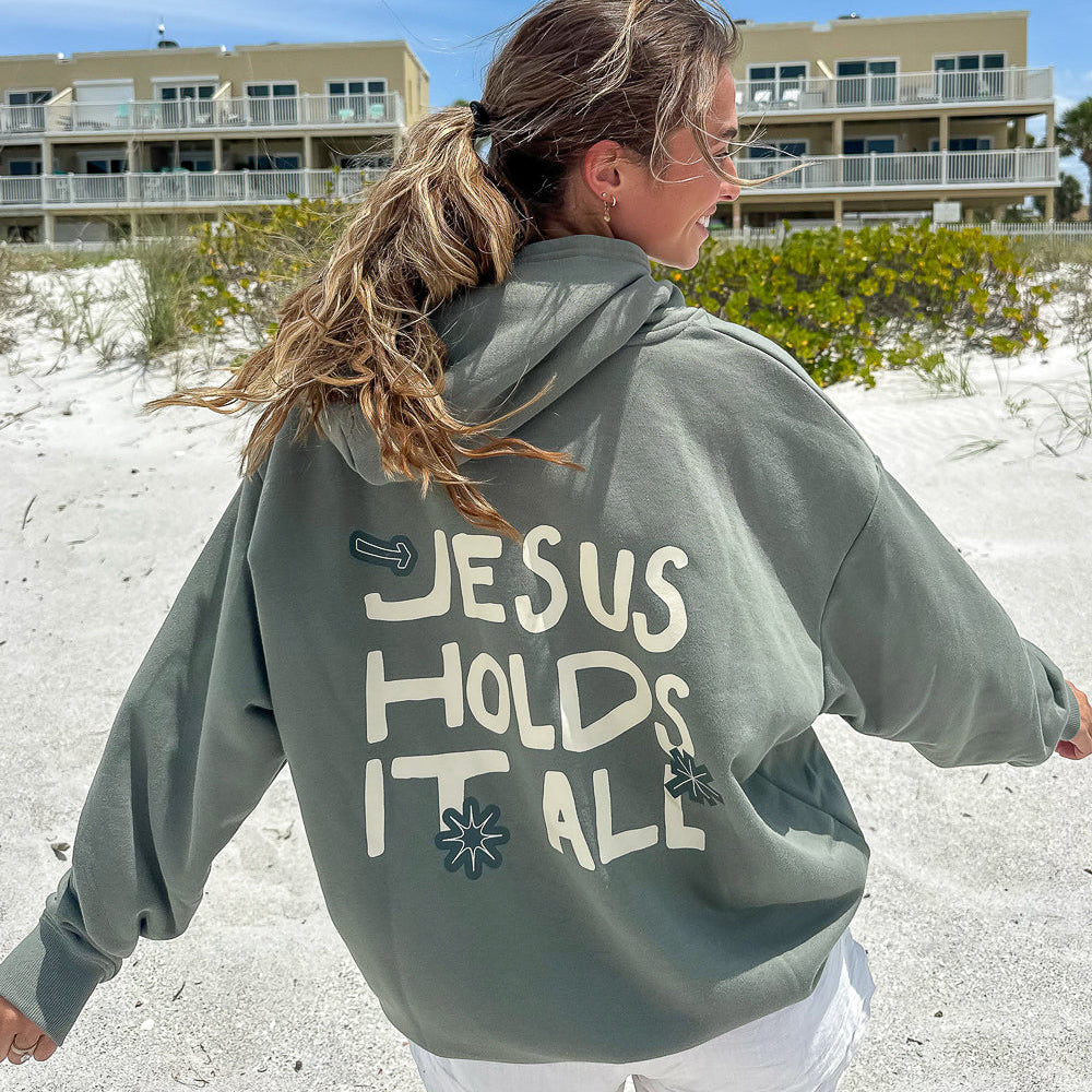 Person wearing a hoodie with 'Jesus Holds It All' text on a beach.