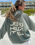 Person wearing a hoodie with 'Jesus Holds It All' text on a beach.