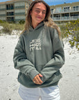 Person wearing a hoodie with 'Jesus Holds It All' text on a beach.