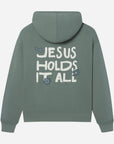 Green hoodie with 'Jesus Holds It All' text on the back against a light gray background