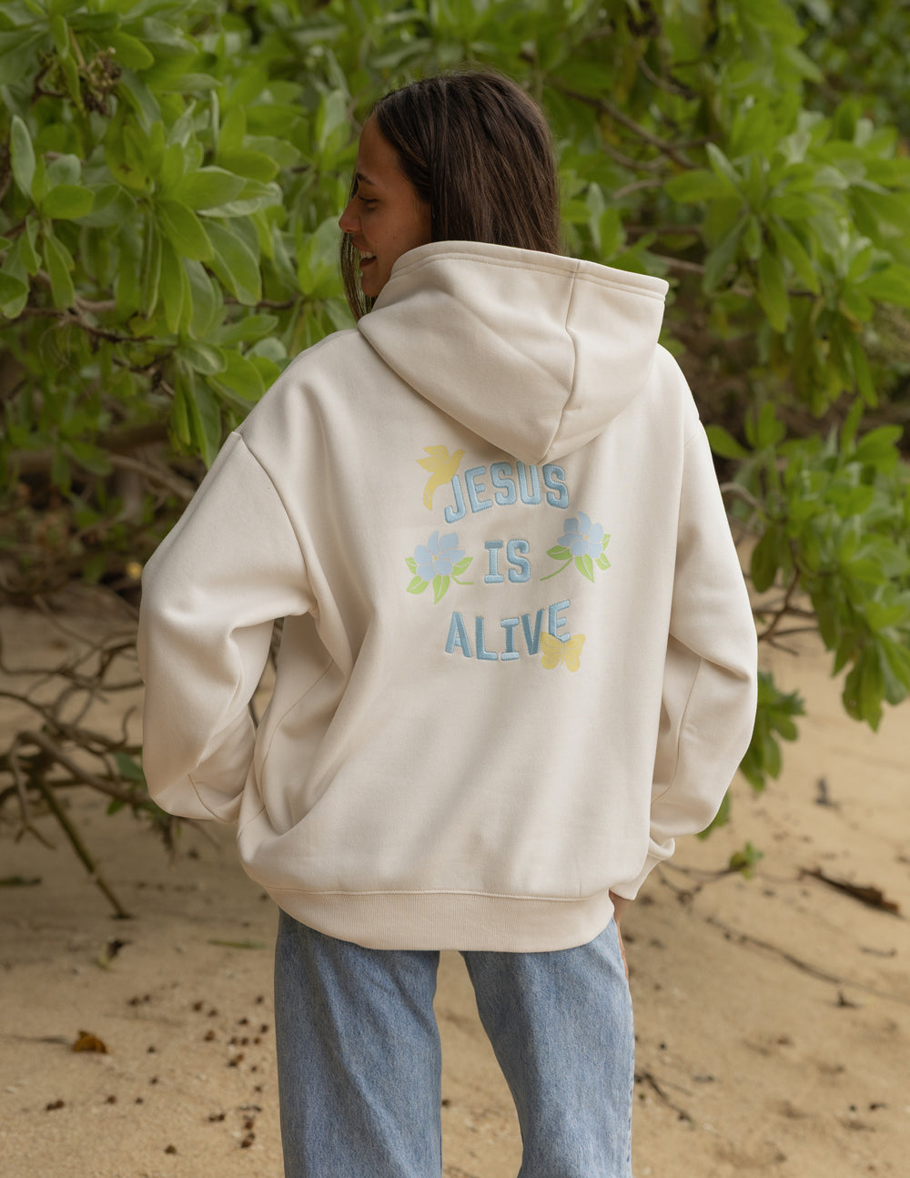 Model wearing the Nimbus Cloud Jesus Is Alive hoodie with pastel lettering and floral accents on the back