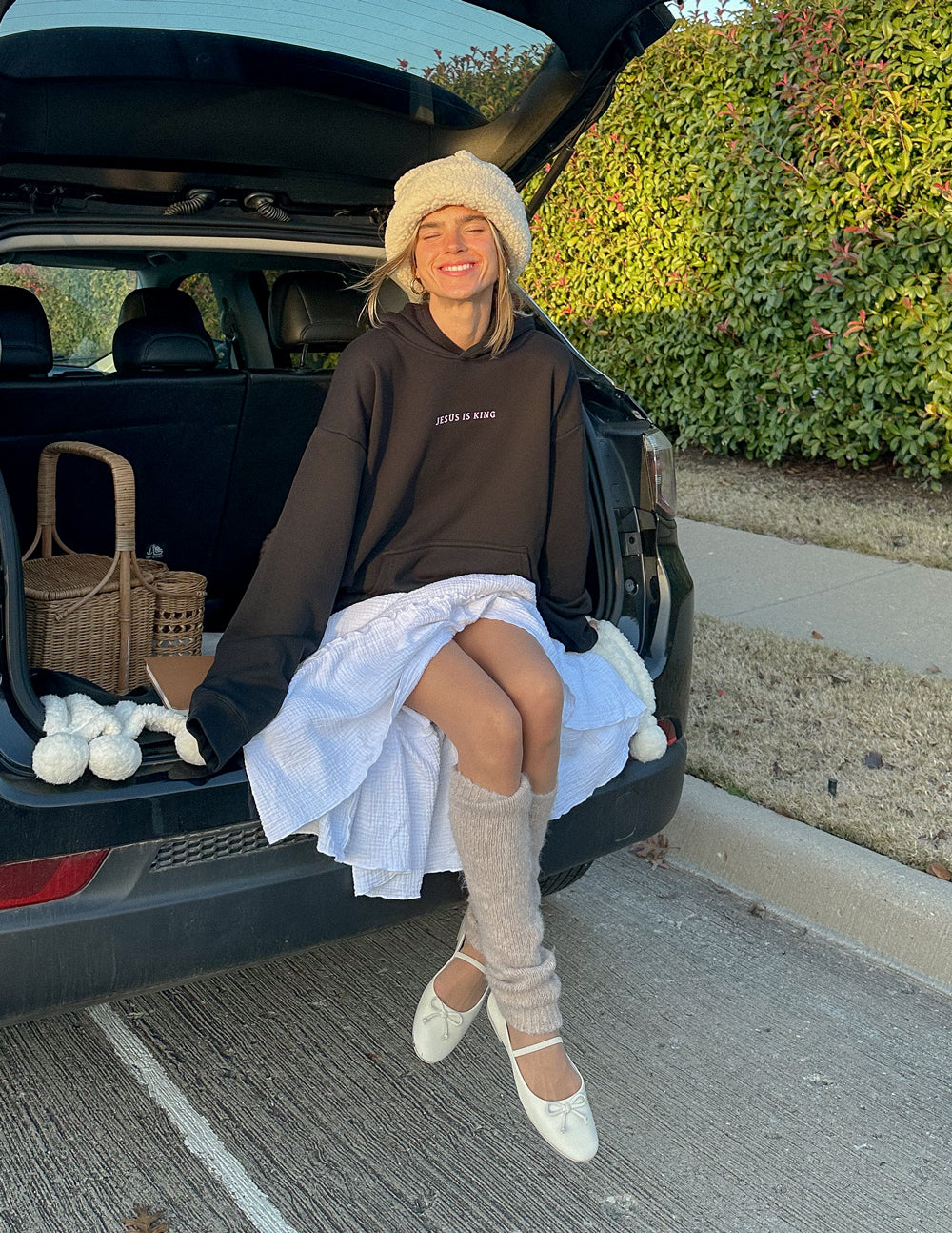 Model seated at an open car trunk wearing the Jesus Is King Black Hoodie with a light-colored skirt