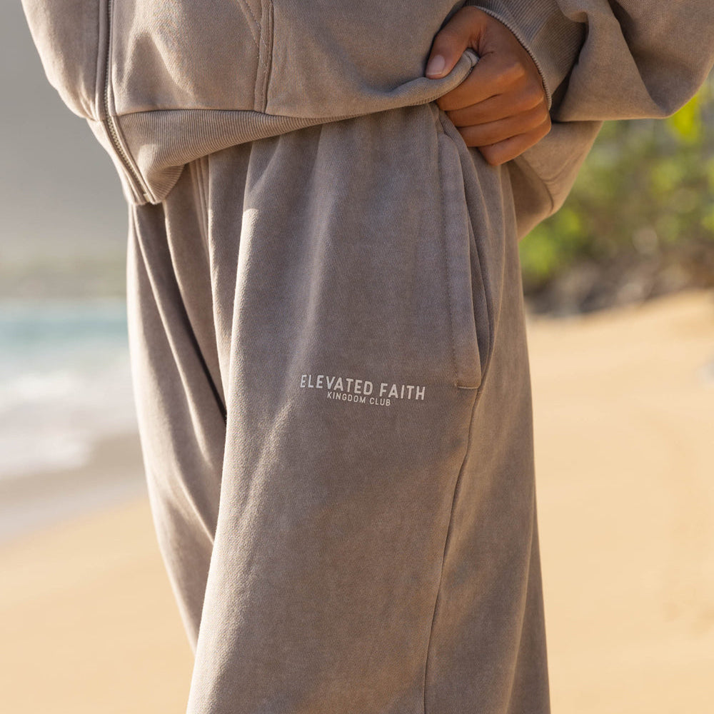 Person wearing a beige hoodie and pants with 'Elevated Faith' logo on a beach.