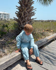 Child seated outdoors beside a palm tree wearing the Air Blue Kingdom Kids Hoodie