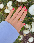 Lilac Enamel Cross Ring worn on a hand with pink nails and a blue sleeve beside white flowers