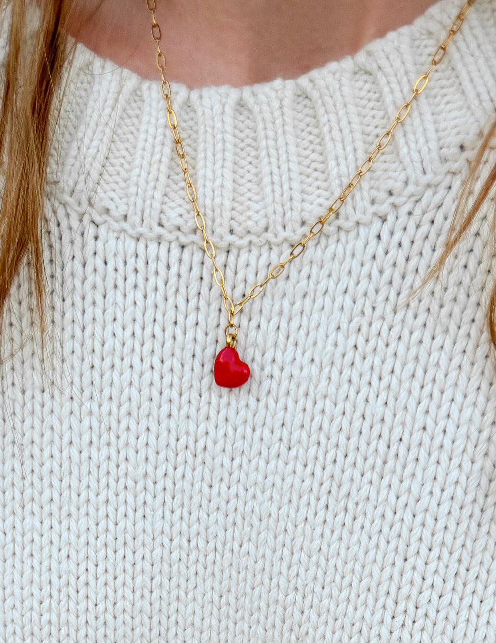 Close-up of the red heart pendant and gold-tone paperclip chain worn with a cream sweater