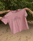 Ash Rose Love Your Neighbor Heavyweight Tee held up outdoors showing the front lettering and short sleeves