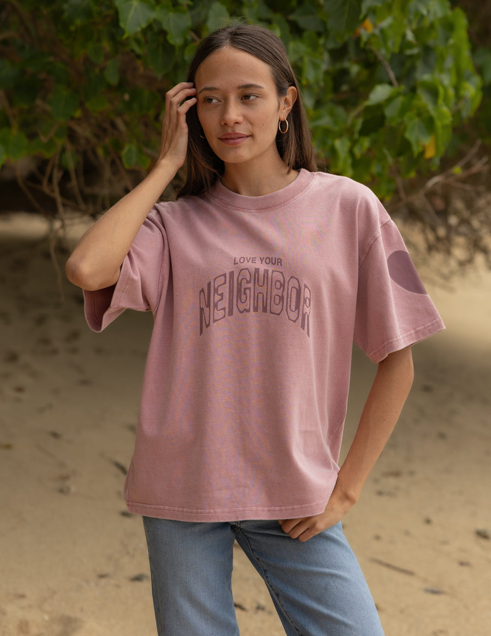 Front view of the Ash Rose Love Your Neighbor tee styled with jeans against green foliage