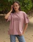Front view of the Ash Rose Love Your Neighbor tee styled with jeans against green foliage