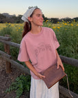 Model wearing the Ash Rose Love Your Neighbor Heavyweight Tee with a white skirt and holding a Bible beside a wildflower field