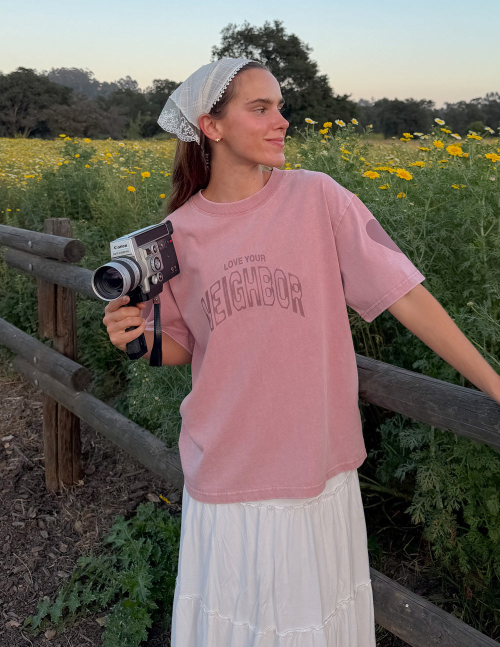 Model wearing the Ash Rose Love Your Neighbor Heavyweight Tee beside a wooden fence and yellow wildflowers