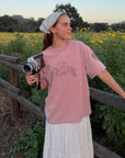 Model wearing the Ash Rose Love Your Neighbor Heavyweight Tee beside a wooden fence and yellow wildflowers