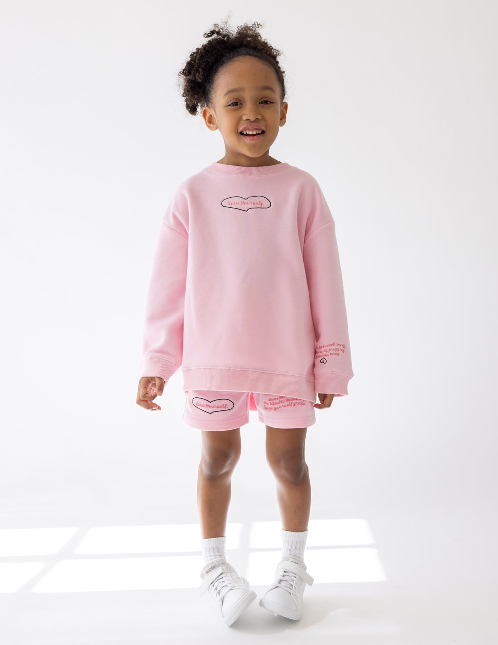 Child wearing the Love Yourself Pink Cherub crewneck sweatshirt with coordinating shorts and white sneakers.