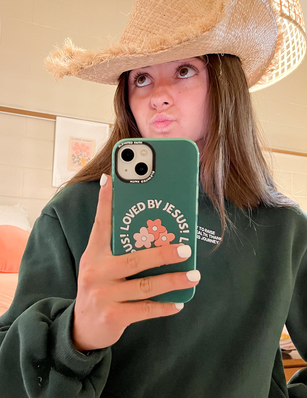 Woman wearing a straw cowboy hat and green sweatshirt holding the Loved by Jesus floral iPhone case for a mirror selfie