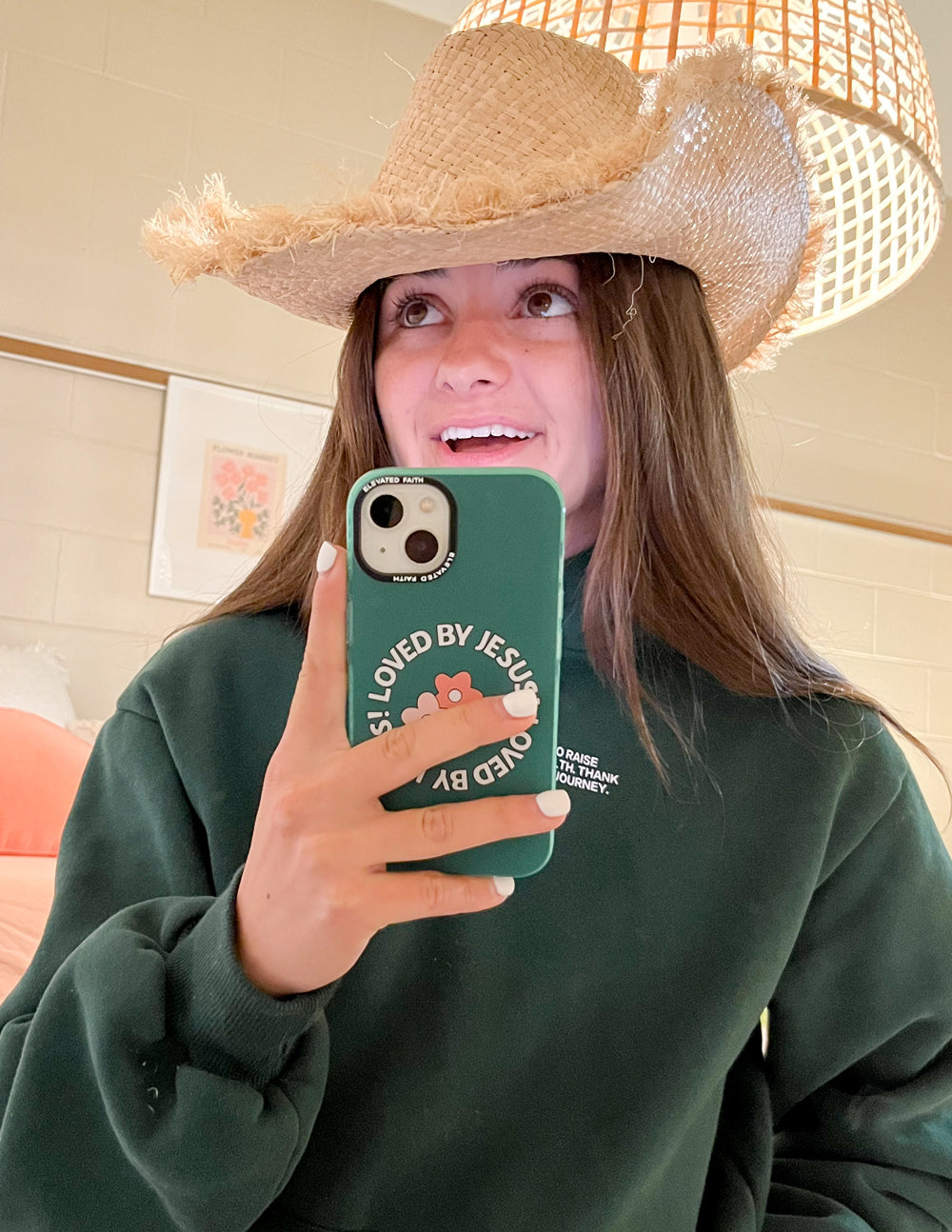 Smiling woman in a straw cowboy hat showing the green Loved by Jesus floral iPhone case in a mirror