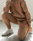 Person wearing a brown hoodie and pants with red text, sitting on the floor.