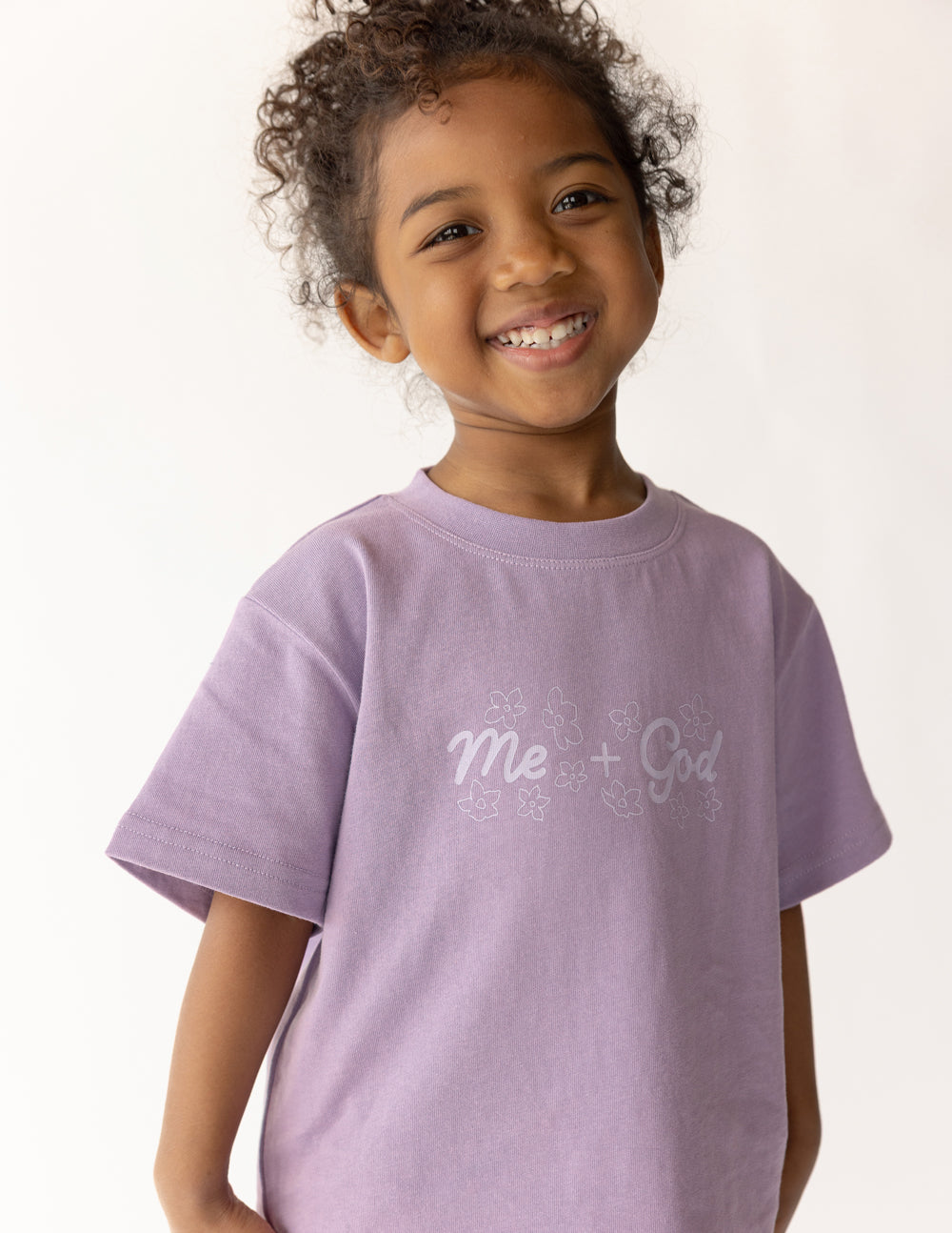 Smiling child wearing the Quiet Violet Me Plus God Kids Tee with light front lettering