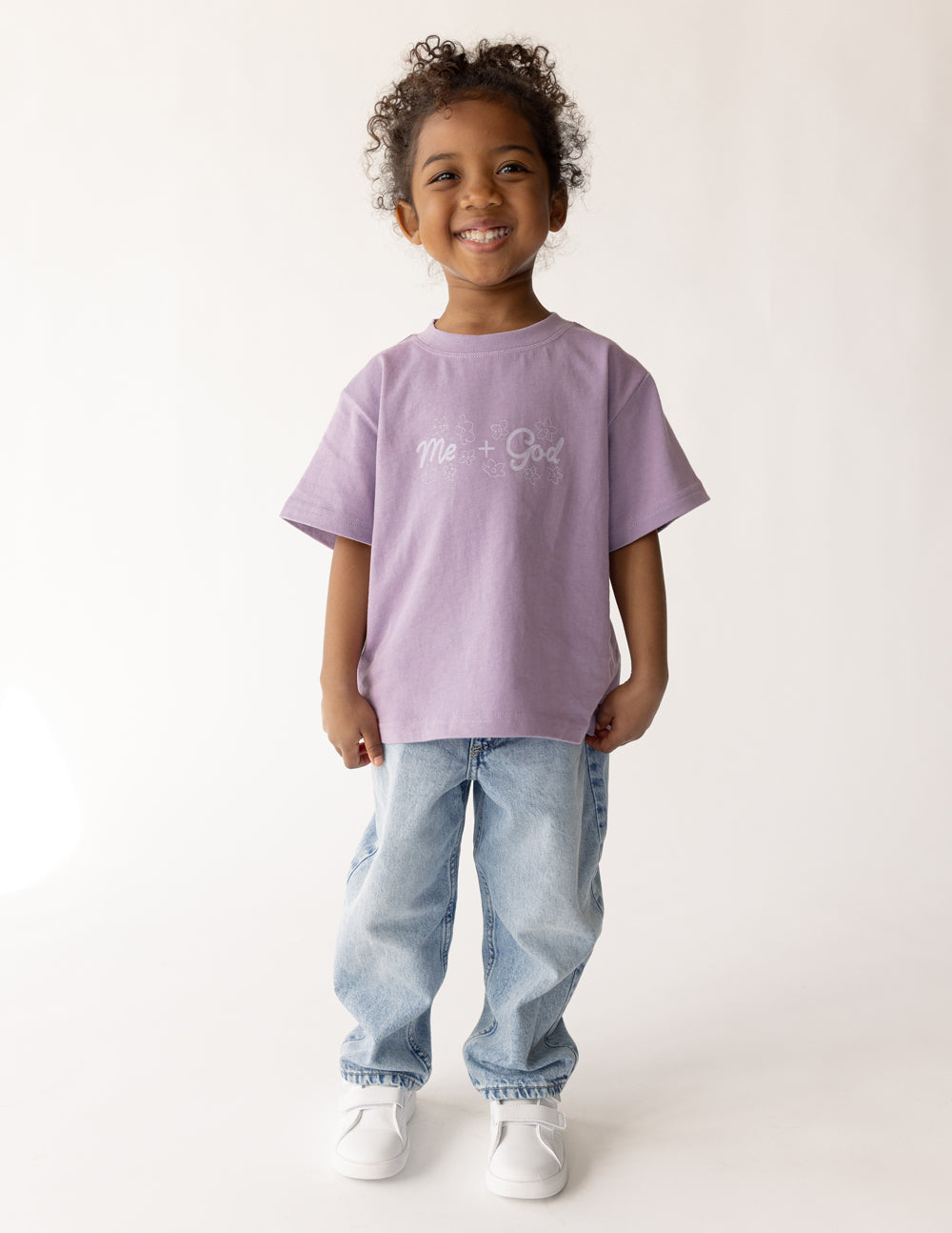 Child modeling the oversized Quiet Violet Me Plus God Kids Tee with jeans and white sneakers