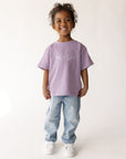 Child modeling the oversized Quiet Violet Me Plus God Kids Tee with jeans and white sneakers