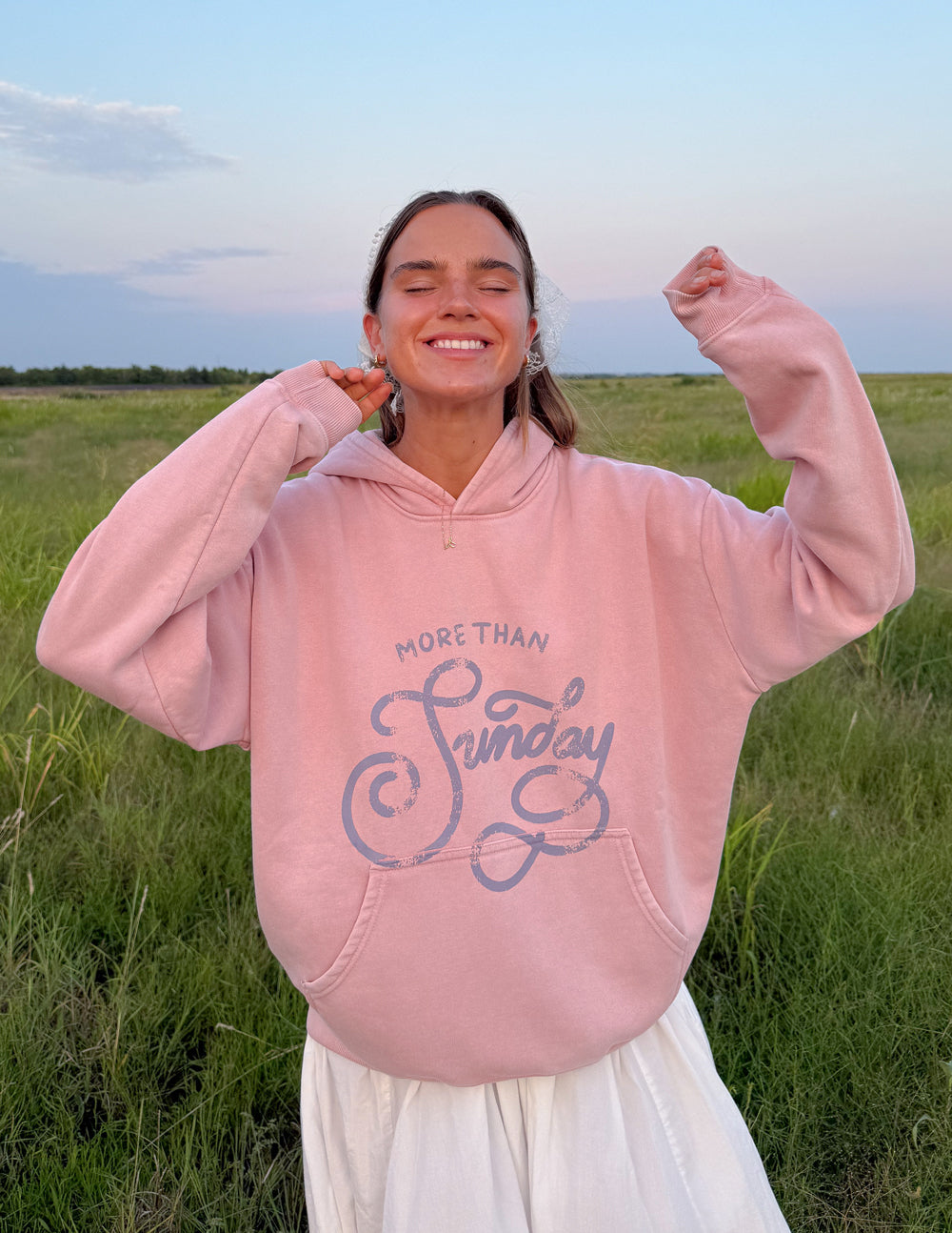 Model outdoors wearing the More Than Sunday Zephyr Pink Hoodie with blue script lettering