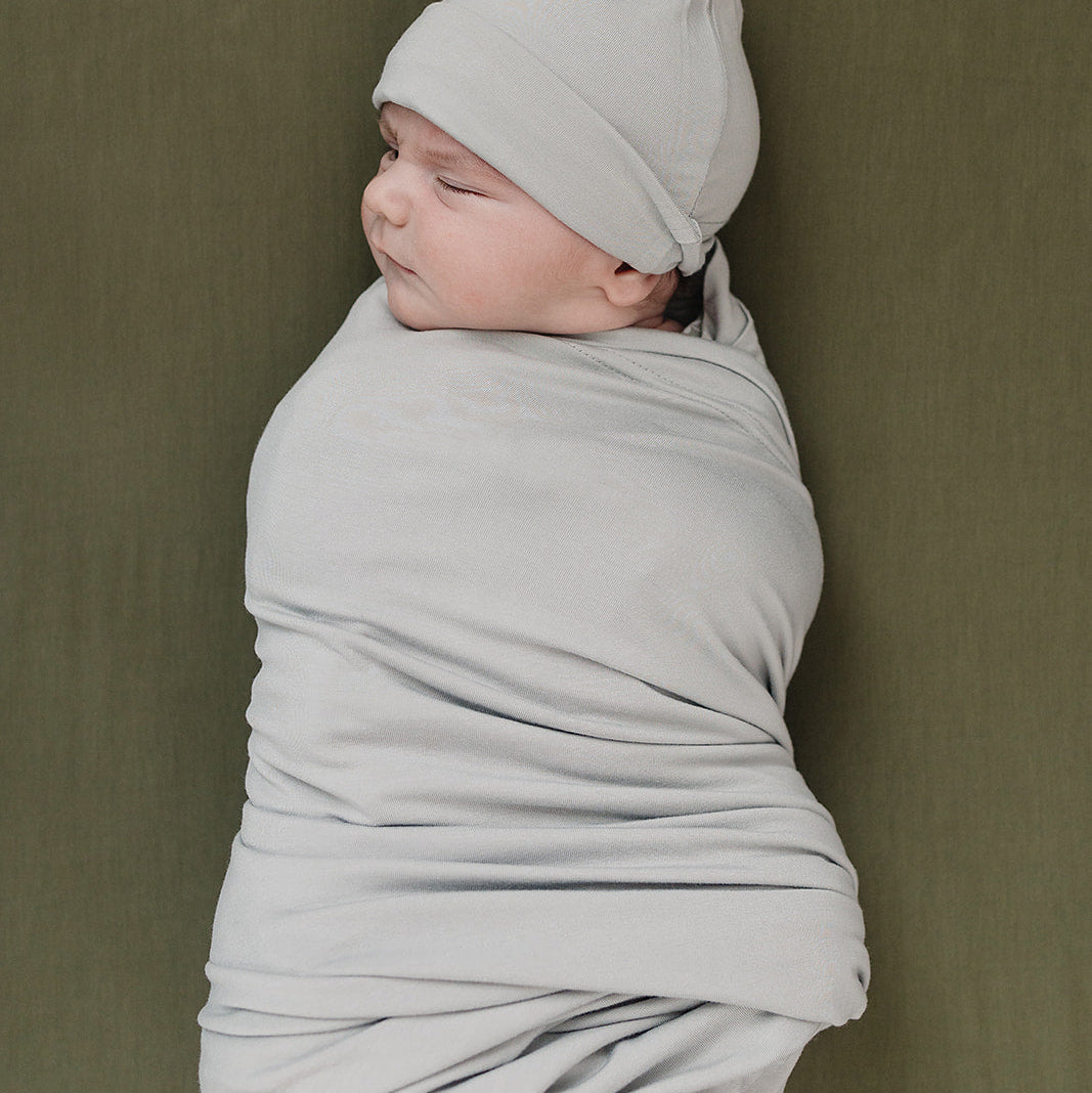 Baby wrapped in a light gray swaddle and cap against a green background
