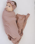 Newborn baby wrapped in a brown blanket with a matching headband, lying on a white surface.