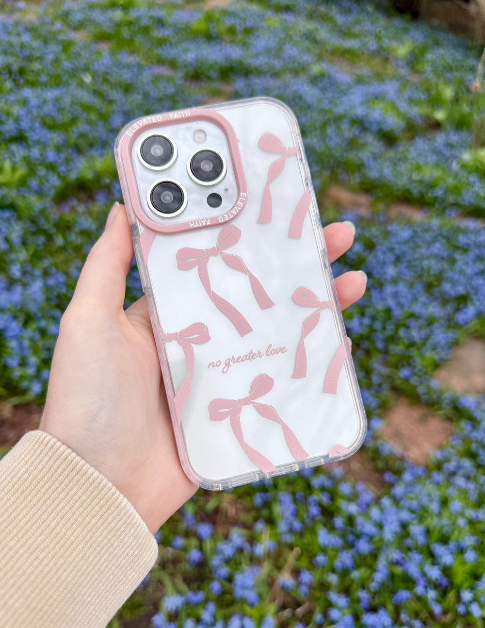 Hand holding an iPhone in the pink bow No Greater Love case above small blue flowers.