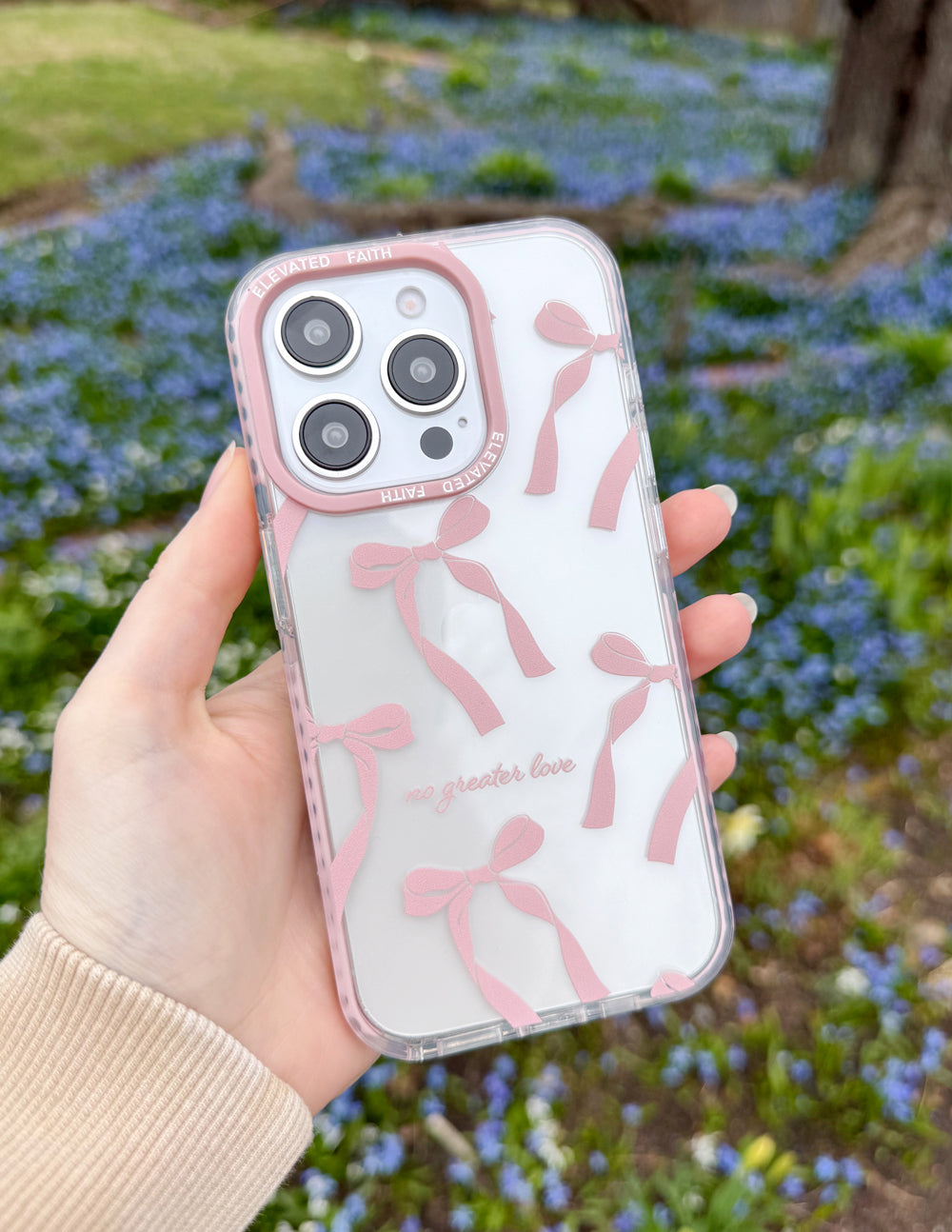 Angled view of the No Greater Love pink bow iPhone case held outdoors above blue flowers.