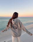 Woman wearing the Not of This World Hoodie beside the ocean at sunset, showing the back design.