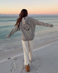 Woman walking along the beach with arms extended in the Heather Greige Not of This World Hoodie.