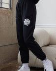 Person wearing black sweatpants with text and white sneakers in a room.