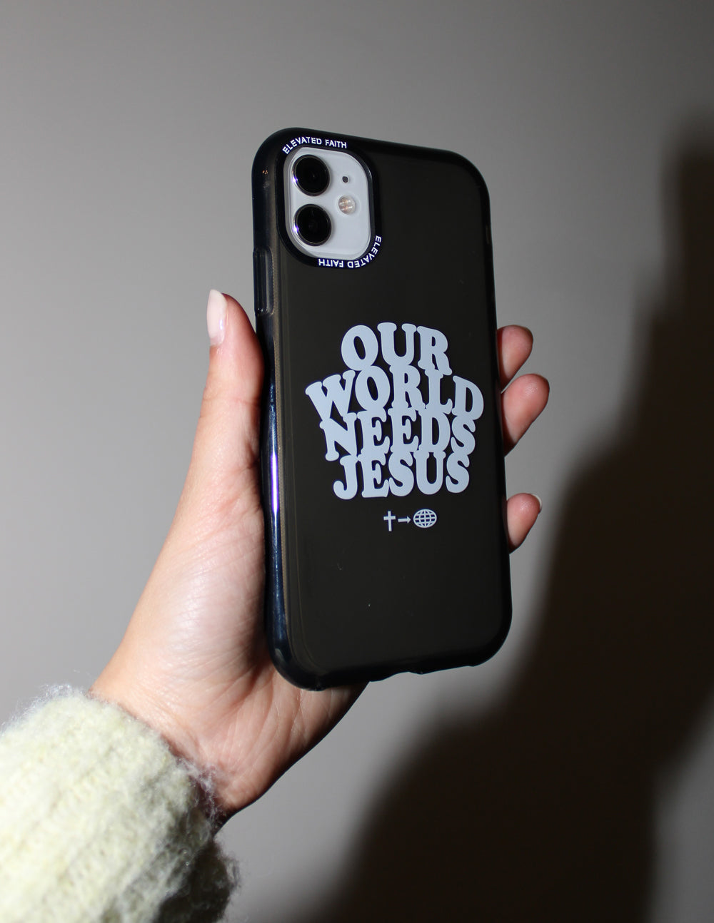 Hand holding a dark Our World Needs Jesus iPhone case against a softly lit wall